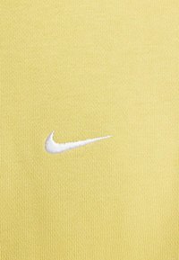 Tessuto atletico giallo con superficie testurizzata, caratterizzato da un logo Nike Swoosh ricamato in bianco posizionato sulla sinistra.