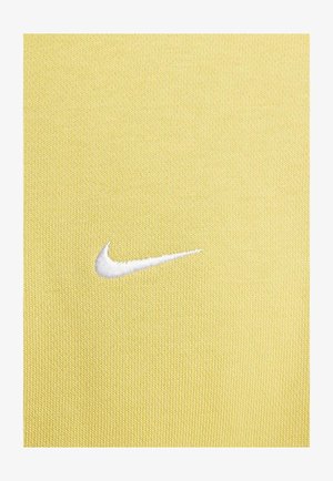 Geel atletisch stof met een gestructureerd oppervlak, voorzien van een witte geborduurde Nike Swoosh-logo aan de linkerkant.