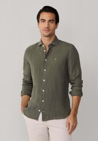 Man draagt een olijfgroen overhemd met opgerolde mouwen en lichtbeige broek, staand tegen een effen lichte achtergrond.