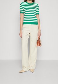 Top tricotato a righe verdi e bianche con maniche corte, abbinato a pantaloni larghi di colore crema. Porta una piccola borsa a tracolla arancione lucida.