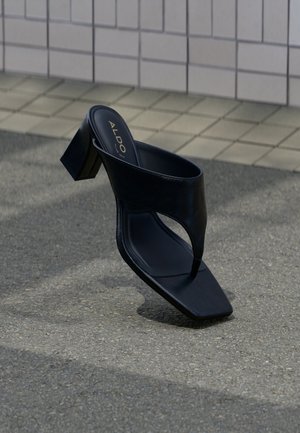 Sandale à talon noire avec bout carré et large bride, posée sur un bitume gris près d'un mur carrelé.