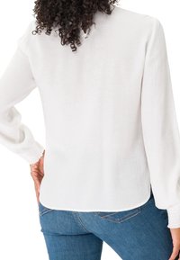 Femme aux cheveux bouclés portant un chemisier blanc texturé à manches longues et un jean bleu, vue de dos avec une main sur la hanche.