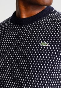 Pull en tricot bleu marine avec un motif texturé de petits points blancs. Doté d'un col rond et d'un logo brodé vert sur la poitrine.