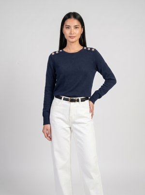 Frau mit langen schwarzen Haaren, die einen dunkelblauen Pullover mit Schulterknöpfen, weiße hoch taillierte Hose und einen schwarzen Gürtel trägt, steht mit der Hand in der Hüfte.
