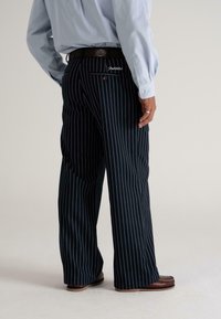 Man in een zwart gestreepte, brede broek, lichtblauw shirt, een bruine riem en bruine schoenen, staande met ontspannen handen langs zijn zij.