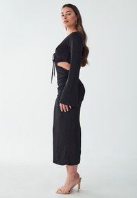 Black long-sleeve dress with a fitted design, cut-out detail, and ruched waist. Soft fabric with a smooth texture and a straight skirt. Clear heeled sandals.
