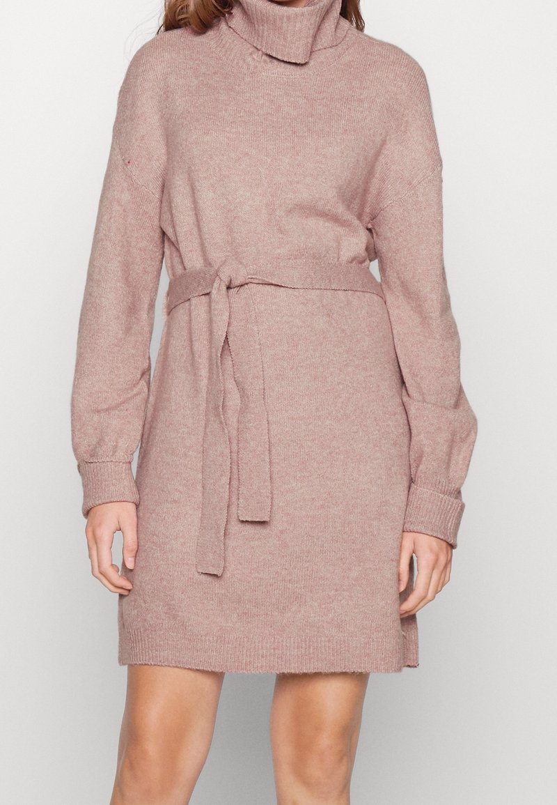 Femme portant une robe à manches longues en maille rose pâle avec un col roulé et une ceinture nouée à la taille, debout devant un fond uni.