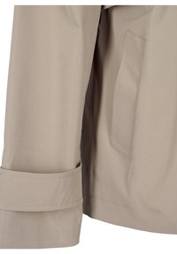 Gros plan sur la manche beige du manteau et le côté avec une poche cousue et un détail de sangle près du poignet sur un tissu lisse.