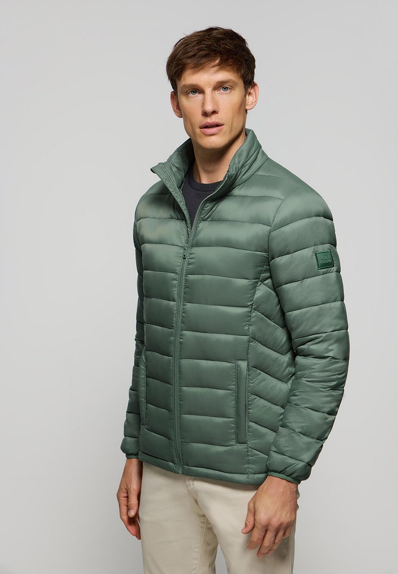 Chaqueta verde acolchada con cremallera frontal, cuello alzado y bolsillos laterales. Cuenta con un parche de marca en la manga izquierda. Textura suave.
