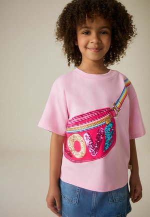 Lapsi, jolla on kiharat hiukset, pukeutuneena vaaleanpunaiseen paitaan, jossa on värikäs paljetein koristeltu "LOVE"-vyölaukku, ja farkkushortsit, yksiväristä taustaa vasten.