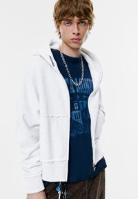 Fiatal személy fehér cipzáros kapucnis felsőt visel tengerészkék grafikai pólóval, ezüst láncra akasztott nyaklánccal, egy sima fehér háttéren.