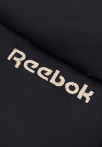 Logo Reebok w beżowym haftowaniu na czarnej, gładkiej powierzchni materiału, prezentujący prostokątny wzór o miękkiej fakturze i czystych liniach.