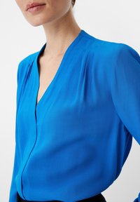 Blusa blu a maniche lunghe con scollo a V, morbida al tatto, con pieghe sottili sulle spalle e una silhouette aderente.