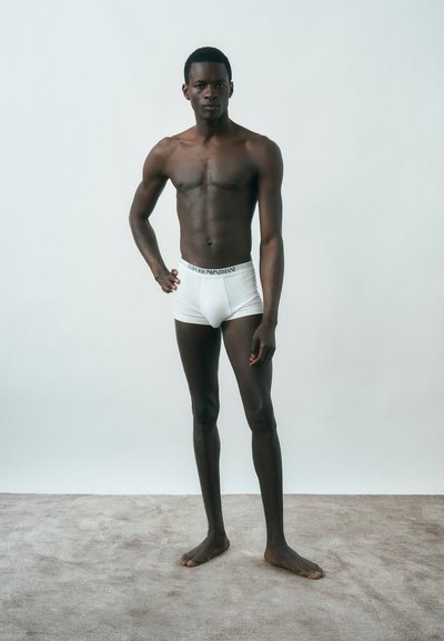 Ψηλός άνδρας χωρίς πουκάμισο στέκεται ξυπόλυτος σε χαλί φορώντας λευκά εσώρουχα Emporio Armani με φόντο απλό και φωτεινό.