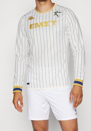 Homme portant un maillot de sport blanc à manches longues avec de fines rayures bleu marine, des accents dorés et un short blanc avec un logo noir sur la jambe.