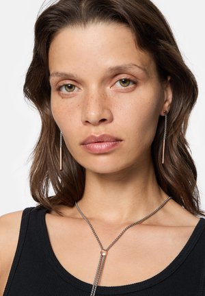 Ohrringe aus Roségold im Tropfenstil mit langen, schlanken Ketten und zylindrischen Akzenten. Das Model trägt ein schwarzes Tanktop und eine geschichtete Silberkette.