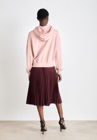Sudadera con capucha rosa combinada con una falda de tablas burdeos hasta la rodilla, que presenta una tela texturizada y zapatos de tacón alto negros.