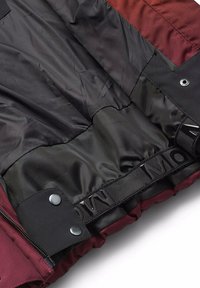 Interno di un giubbotto imbottito rosso e nero, caratterizzato da una morbida fodera nera, una fascia con logo impresso e chiusure a scatto vicino alla base.
