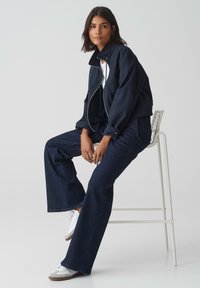 Marineblauwe cropped jas met hoge kraag en zilveren rits, gecombineerd met donkere flared jeans en witte sneakers, zittend op een witte kruk.