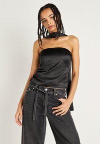 Top sem mangas em cetim preto com corte justo e detalhe de nó. Combinado com jeans pretos de cintura alta e corte solto, com um cinto na cintura.