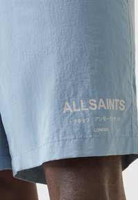 Pantalones cortos azul claro de tela arrugada, con un texto de logo blanco y caracteres japoneses adicionales cerca del dobladillo.