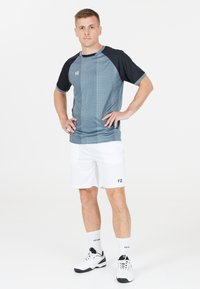 Man står med händerna på höfterna, iklädd en blå och svart sporttröja, vita shorts, vita strumpor och vita sportskor framför en vit bakgrund.