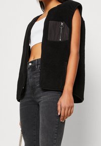 Mustavilla fleece-vestillä, jossa on hihattomuus, vetoketjullinen rintatasku ja kontrastiväriset reunat. Yllä valkoinen crop-top ja tummat farkut.