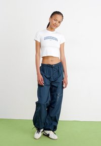 Bílý zkrácený tričko s nápisem "BOHYNĚ"; tmavě námořnické volné kalhoty; bílé sneakers s černými akcenty; jednoduché pozadí, zelená podlaha.