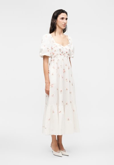 Γυναίκα στέκεται φορώντας ένα λευκό φόρεμα μέχρι τους αστραγάλους με φουσκωτά μανίκια και μικρά ροζ λουλουδάτα σχέδια, σε συνδυασμό με λευκά μυτερά παπούτσια.