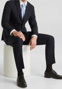 Completo blu notte con vestibilità slim, cravatta azzurra chiara, camicia a righe bianche, scarpe nere e calzini neri a fantasia, seduto su un piedistallo bianco.