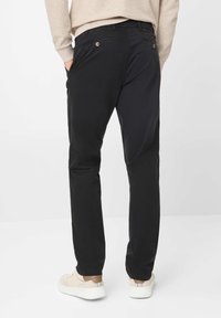 Pantaloni neri con una texture liscia, caratterizzati da tasche posteriori e dettagli in bottone, abbinati a sneakers di colore chiaro.