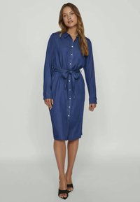 Ženska stoji v modri obleki do kolen s dolgimi rokavi, gumbom ob sprednjem delu, z obrobo za pas in črnimi salonarji brez prstov.