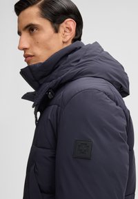 Mann in einer dunklen gepolsterten Jacke mit hohem Kragen und Kapuze, die ein schwarzes Patch mit einem Kreuzsymbol am oberen Ärmel zeigt.
