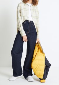 Chemise à carreaux claire à boutons, t-shirt blanc, jean en denim foncé taille haute, et une doudoune noire et jaune, tenue dans une main.