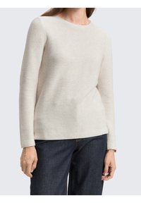Femme portant un pull en maille côtelée crème à manches longues, associé à un jean bleu foncé, sur un fond clair uniforme.