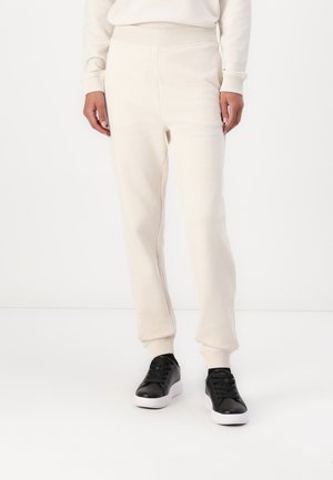 Henkilö pukeutunut kermaisiin collegehousuihin ja vastaavaan yläosaan, mustat lenkkarit jalassa, seisoo yksivärisen valkoisen taustan edessä.