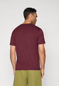 Ein Mann, der von hinten zu sehen ist, trägt ein schlichtes, kastanienbraunes T-Shirt mit kurzen Ärmeln und olivgrüne Shorts vor einem neutralen Hintergrund.