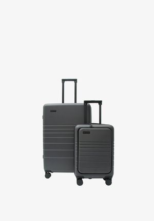 Dwie twarde walizki w ciemnoszarym kolorze z wysuniętymi teleskopowymi rączkami i czterema kółkami, jedna duża i jedna średniej wielkości, na białym tle.