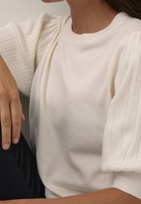 Suéter de punto acanalado de crema clara con mangas abullonadas y cuello redondo. Textura suave, cuerpo ajustado y detalles verticales sutiles en las mangas.