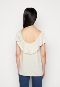 Lys beige off-shoulder top med en flæset neckline og ryg, lavet af et blødt stof. Sammen med blå jeans.