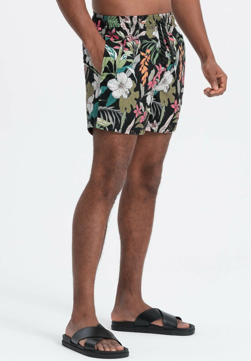 Homme portant un short noir à motif floral et des sandales noires à enfiler, debout avec une main dans la poche et l'autre détendue.