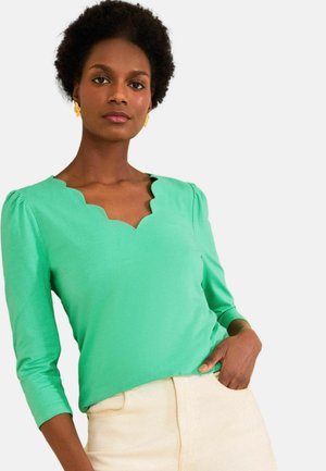 Frau mit kurzen lockigen Haaren, die gelbe Blumenohrringe, ein grünes Top mit gewelltem Ausschnitt und cremefarbene Hosen trägt, mit einer Hand in der Tasche.