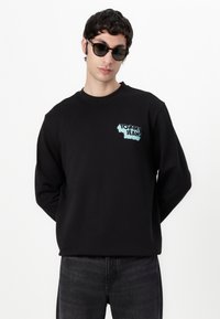 Zwarte sweatshirt van zacht materiaal, met een blauw logo op de borst. Gepaard met donkere jeans en ronde zonnebril.