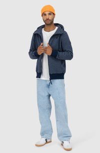 Felpa con zip a righe blu navy e bianche, con un ampio cappuccio, indossata sopra una maglietta bianca, abbinata a jeans azzurri chiari e sneaker bianche.