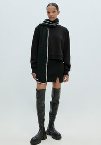 Schwarzes, kurzes Sweatshirt kombiniert mit einem schwarzen, gerippten Minirock mit einem Schlitz vorne, ergänzt durch einen dunkelgrünen Strickschal mit weißen Akzenten und glänzenden schwarzen Stiefeln bis zum Knie.