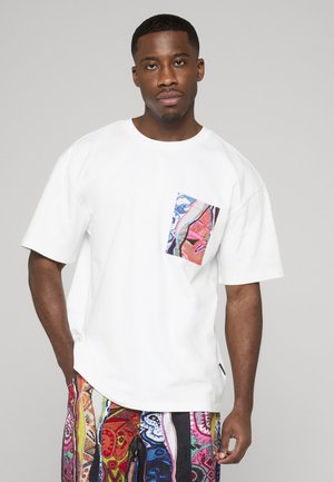 Mand iført hvid oversized t-shirt med farverig abstrakt lomme og matchende flerfarvede mønstrede bukser, stående mod en grå baggrund.