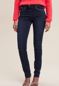 Jean skinny bleu foncé en denim, avec une taille haute, un design à cinq poches et des coutures en contraste. Le mannequin porte des talons avec des lanières.