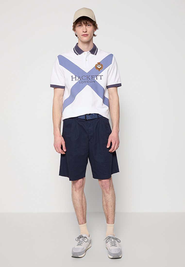 Polo shirt blanc avec des rayures diagonales bleues, logo brodé, short marine, casquette beige et baskets grises avec des accents blancs.
