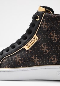 Czarne sneakersy typu high-top z ekoskóry z brązowym nadrukiem logo, złotymi elementami oraz czarnymi sznurowadłami. Biała gumowa podeszwa o teksturowanym wykończeniu.