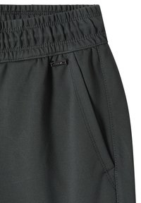 Gros plan sur la ceinture élastique noire et la poche latérale avec un petit logo métallique sur un pantalon ou short en tissu foncé.